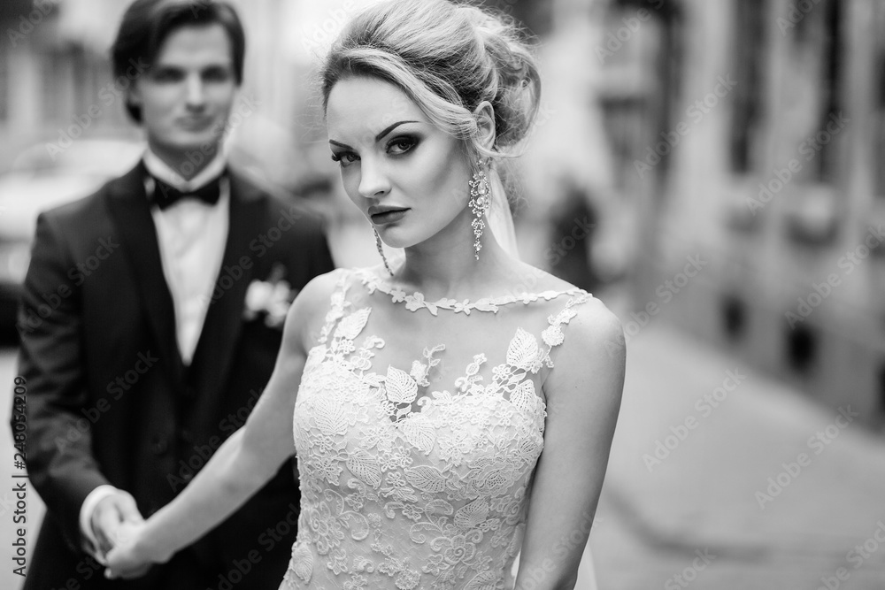 stylish bride and groom walking in city street. luxury wedding couple holding hands at old building in light. romantic sensual moment.  woman looking and man posing, black and white