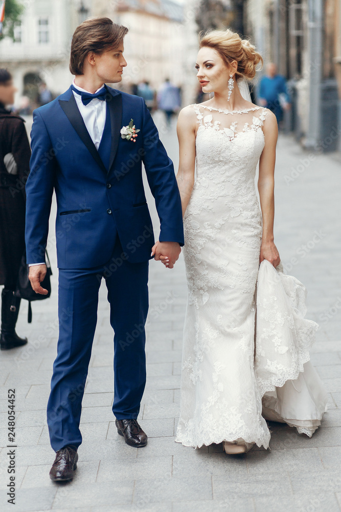 stylish bride and groom walking in city street. happy luxury wedding couple holding hands in light . romantic sensual moment.   man and woman looking at each other