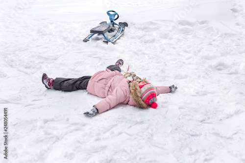 A little girl is lying in snow after falling from his snowmobile, sledge. Children’s going sledding.