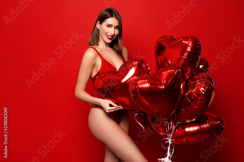 A flirting brunette in red bikini holds a balloon in the shape of a red heart on red background. perfect holiday photo valentines day photo