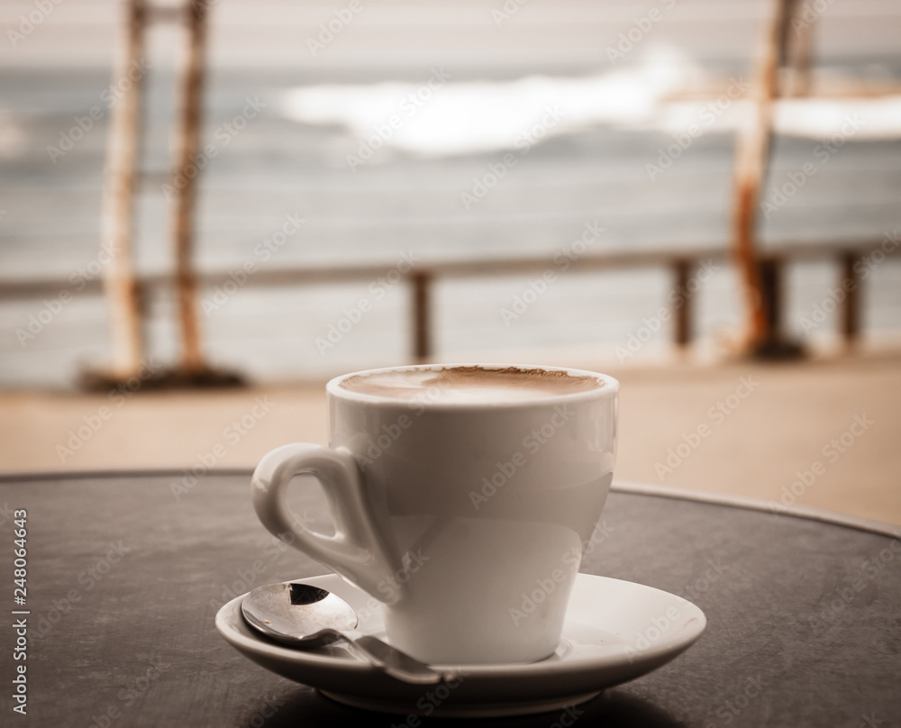 Coffee cup on round table in cafe terrace on the old pier with rusty railing and the view on the sea with waves in cold cloudy day. Loneliness idea. Sepia aged photo.