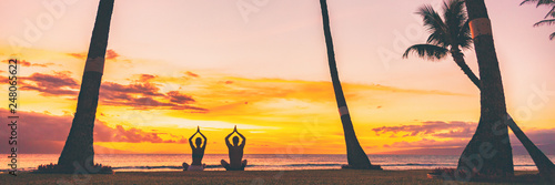Yoga meditation retreat landscape panoramic banner - people meditating on sunset beach praying with hands above head sitting in lotus pose on nature background.