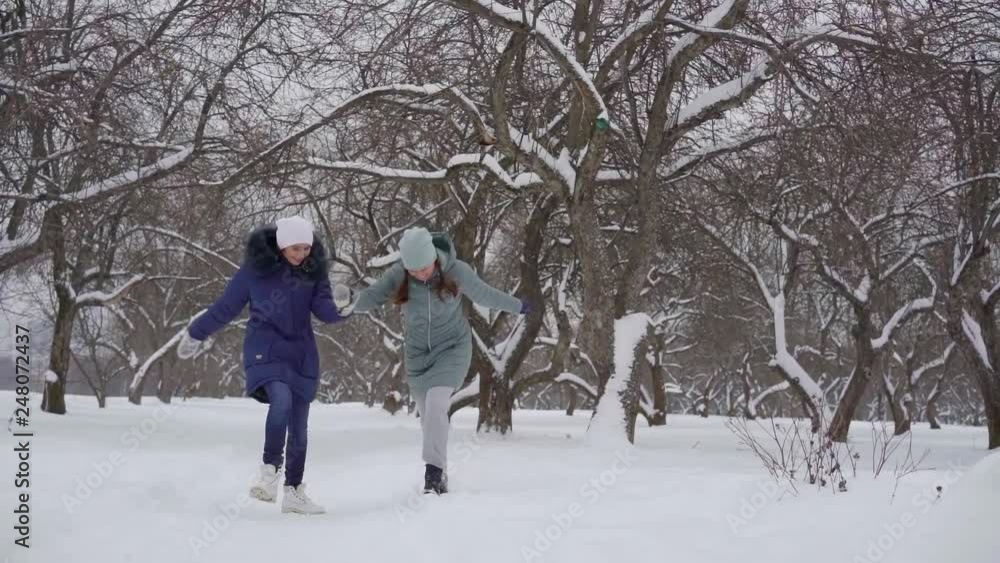 two cheerful adult women are holding hands and leaping over snow in park like little girls, smiling two cheerful adult women are holding hands and leaping over snow in park like little girls, smiling