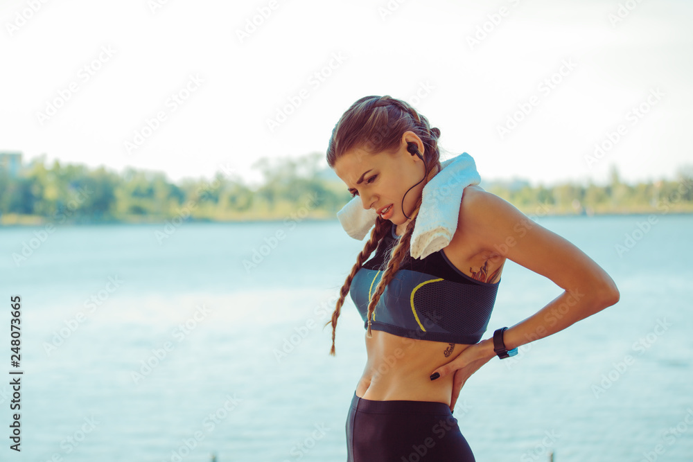 Back pain. Athletic woman in blue black sportswear standing at the seafront rubbing the muscles of her lower back, half body torso portrait. Back pain. Athletic woman in blue black sportswear standing at the seafront rubbing the muscles of her lower back, half body torso portrait.