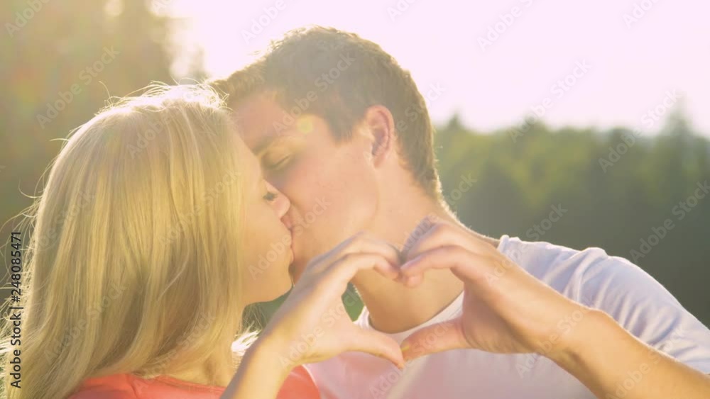 SLOW MOTION, LENS FLARE, PORTRAIT: Cute couple on date in nature kisses gently while making a heart shape with their fingers. Happy boyfriend and girlfriend kiss affectionately on a sunny summer day. SLOW MOTION, LENS FLARE, PORTRAIT: Cute couple on date in nature kisses gently while making a heart shape with their fingers. Happy boyfriend and girlfriend kiss affectionately on a sunny summer day.