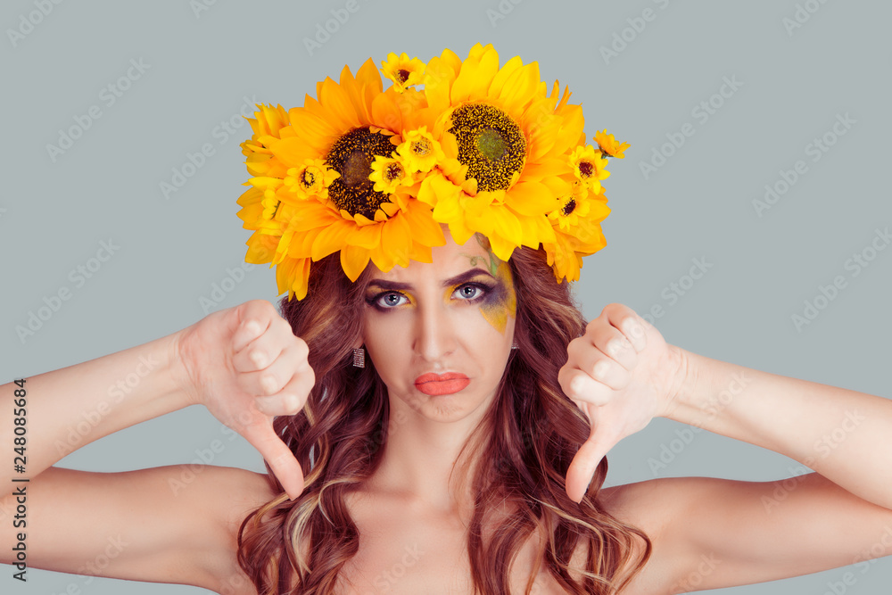 Woman with floral headband with thumbs down isolated over a gray background. Fashion girl with crown from sunflowers on head upset and sad dislikes what she sees.
