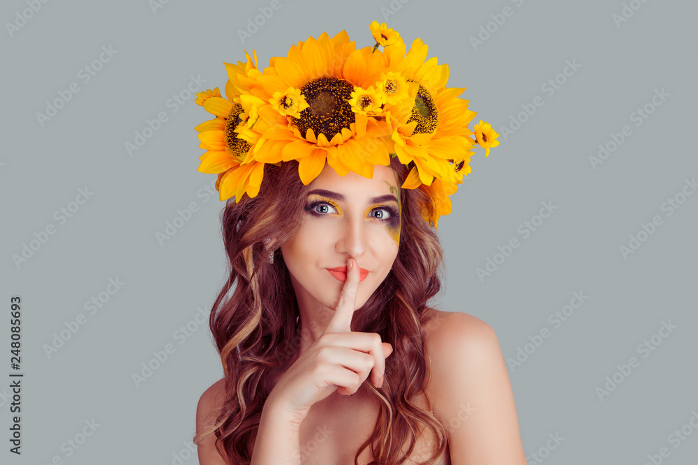 © hbrh - Shh. Young woman with floral headband smiling with a finger to her lips hush sign gesture. Concept for silence. Fashion girl, crown from sunflowers on head isolated on gray background with copyspace.