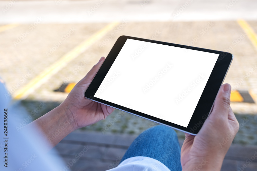 Mockup image of a woman holding black tablet pc with blank white screen horizontally while sitting in the street outdoors