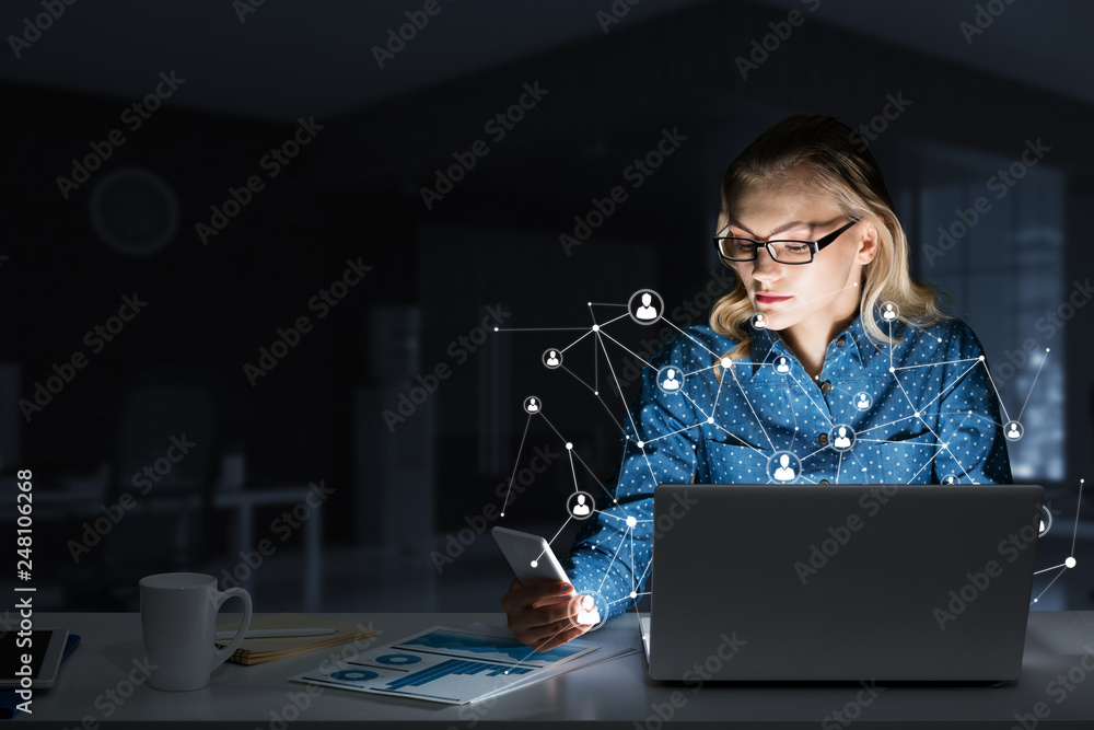 Attractive blonde wearing glasses in dark office using laptop. Mixed media Attractive blonde wearing glasses in dark office using laptop. Mixed media