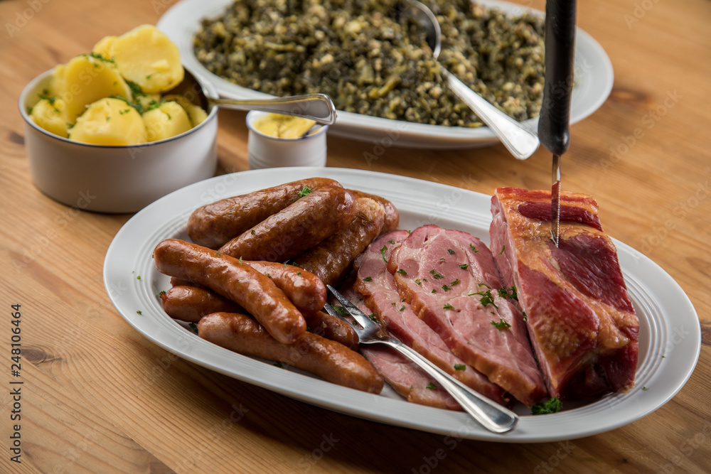 Kassler Nacken, Kohlwurst, Pinkel und Oldenburger Grünkohl auf Platten mit Kartoffeln und Senf auf Holz Tisch Kassler Nacken, Kohlwurst, Pinkel und Oldenburger Grünkohl auf Platten mit Kartoffeln und Senf auf Holz Tisch