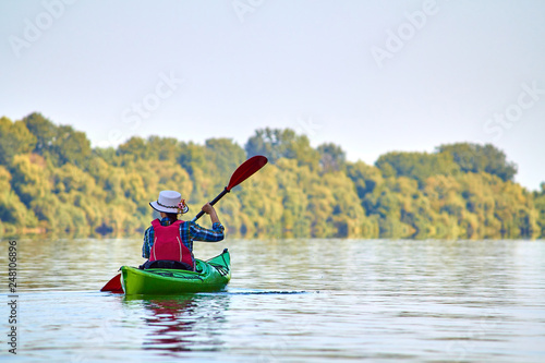 Woman in elegant straw hat canotier paddling green kayak at Danube river. Summer Water Sport