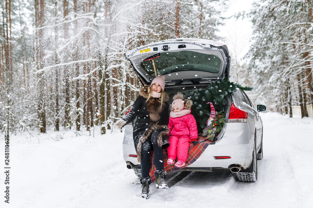 Happy mother with the little daughter and Christmas tree sitting in the car in the winter forest