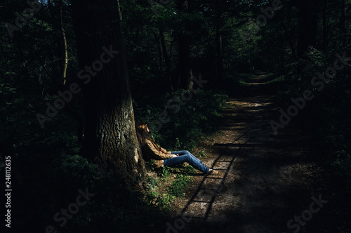 The Young Sad Woman with Hair Covering Her Face Siting Alone Under the Tree Near Footpath with Shadow Lines in the Gloomy Forest