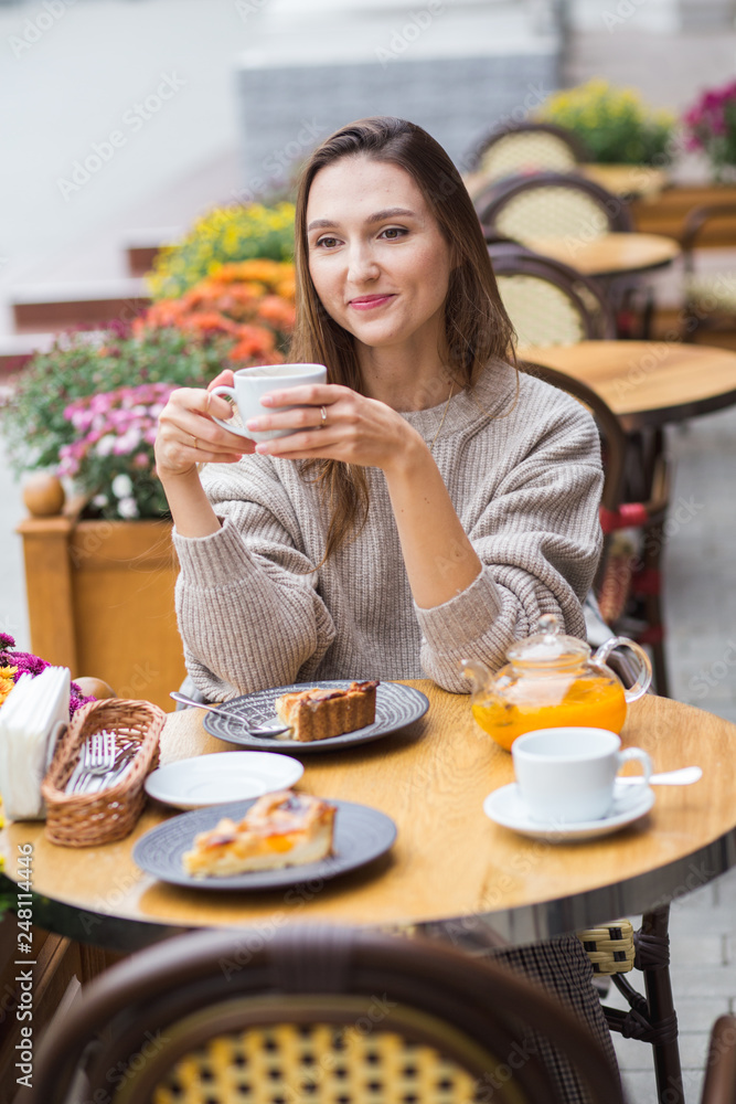 Young attractive smiling woman in gray knitted sweater drinking tea outdoors at the street cafe.