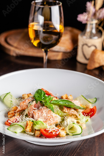 Concept of Italian cuisine. Caesar salad with salmon, lettuce mix, cherry and parmesan cheese. A glass of white wine on the table. Serving dishes in the restaurant. copy space