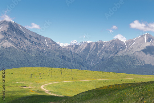 Tranquil scenery with dirt highland road among green meadows and snow capped Caucasus mountains in the background on a sunny summer day. Karachay-Cherkessia, Russia