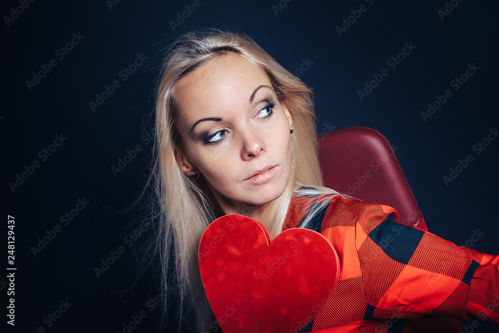 The girl on a black background holds a red Valentine in the form of a heart with a thoughtful look. The girl on a black background holds a red Valentine in the form of a heart with a thoughtful look.