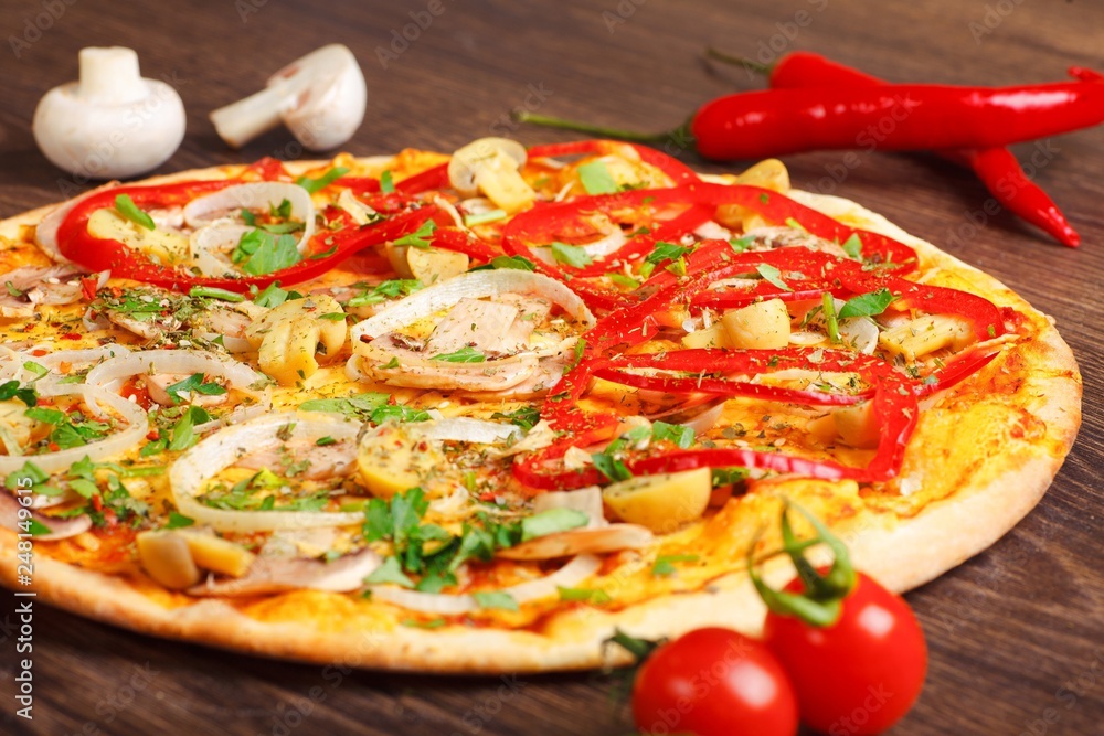 Close up Italian pizza with melted cheese, red tomatoes, pepper, mushrooms, onion and herbs on a brown table decorated by mushrooms, red sweet pepper and cherry tomatoes Close up Italian pizza with melted cheese, red tomatoes, pepper, mushrooms, onion and herbs on a brown table decorated by mushrooms, red sweet pepper and cherry tomatoes