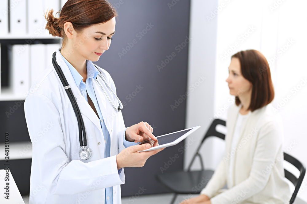 Happy doctor woman at work. Portrait of female physician using tablet computer while standing near reception desk at clinic or emergency hospital. Patient woman sitting at the background. Medicine Happy doctor woman at work. Portrait of female physician using tablet computer while standing near reception desk at clinic or emergency hospital. Patient woman sitting at the background. Medicine