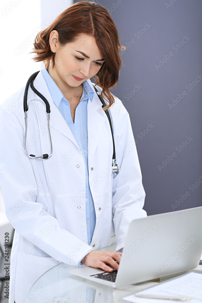 Happy doctor woman at work. Portrait of female physician using laptop computer while standing near reception desk at clinic or emergency hospital. Medicine concept Happy doctor woman at work. Portrait of female physician using laptop computer while standing near reception desk at clinic or emergency hospital. Medicine concept