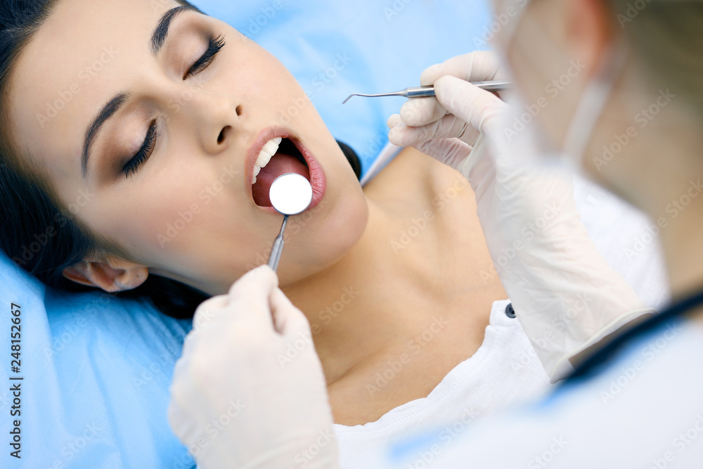 Young female patient visiting dentist office.Beautiful woman with healthy straight white teeth sitting at dental chair with open mouth during oral checkup while doctor working at teeth
