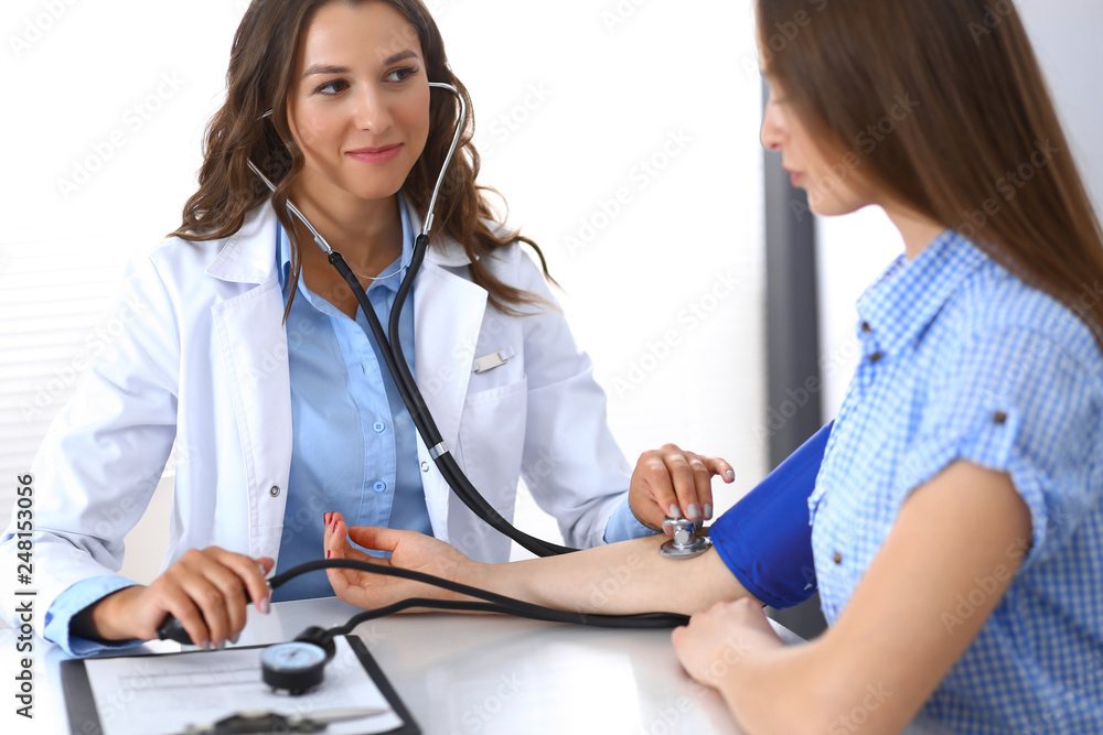 Doctor checking blood pressure of female patient while sitting at the desk in hospital office. Cardiology in medicine and health care concept Doctor checking blood pressure of female patient while sitting at the desk in hospital office. Cardiology in medicine and health care concept