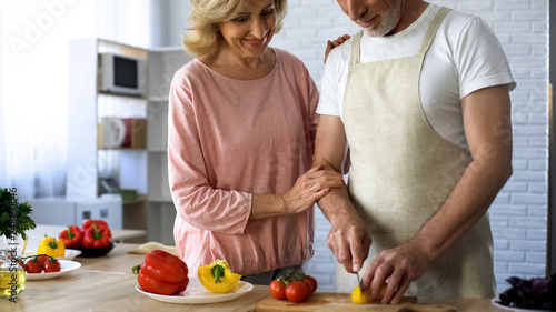 Happy aged wife hugging husband cooking at home, family support, housekeeping
