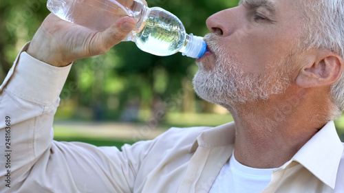 Fotografija Mature man drinking water from bottle in park, maintaining water balance
