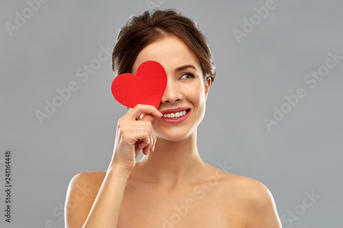 valentine's day, beauty and people concept - happy smiling beautiful young woman covering one eye by red heart over grey background