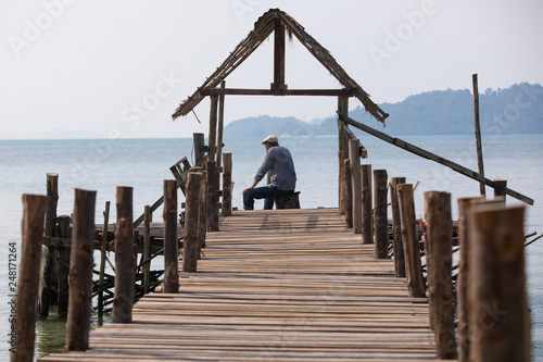 Sad old man in a cap sitting on the pier and looking at the sea, the view from the back