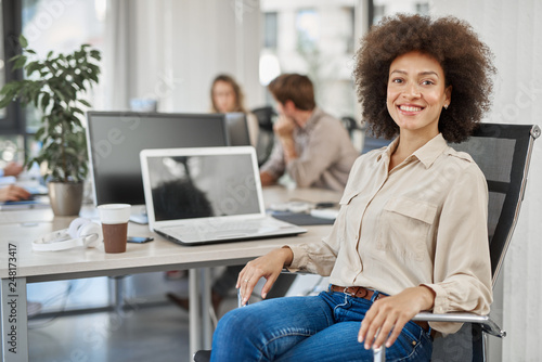 Smiling mixed race CEO sitting at office and looking at camera. In background employees working.