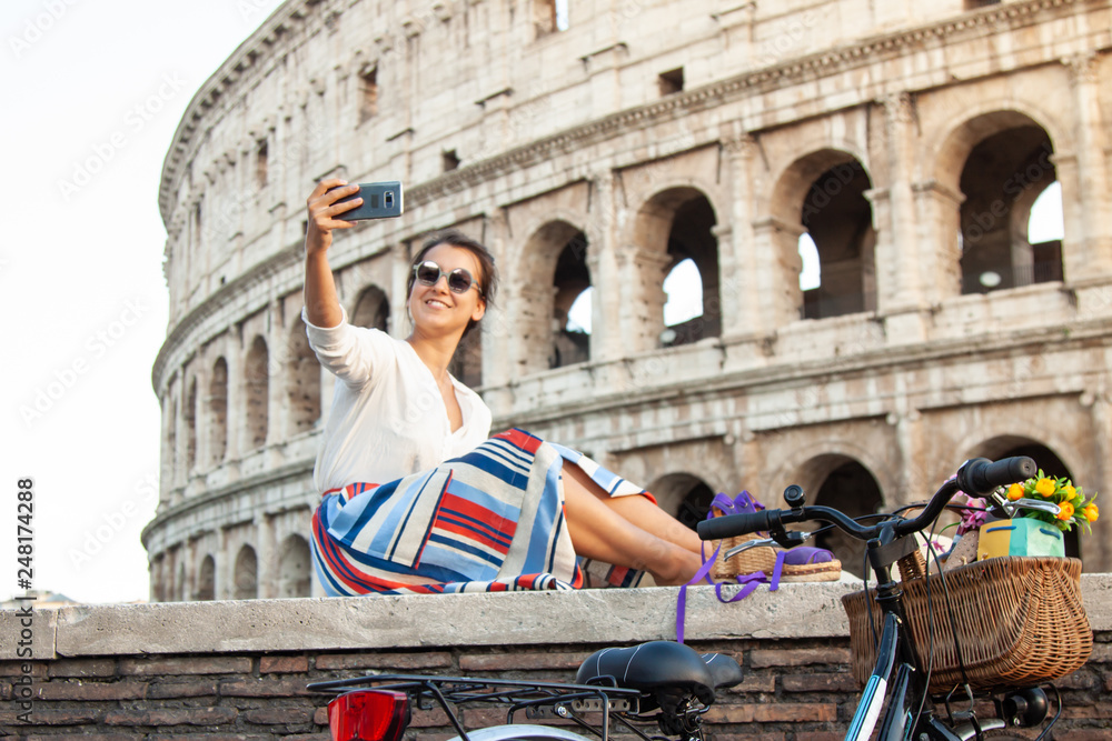 Beautiful young woman taking selfie pictures with smartphone sitting in front of colosseum in Rome at sunset.