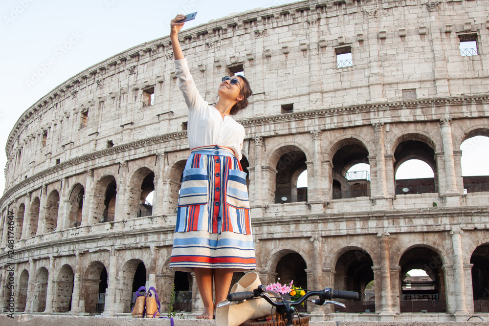 Beautiful young woman taking selfie pictures with smartphone standing in front of colosseum in Rome at sunset.