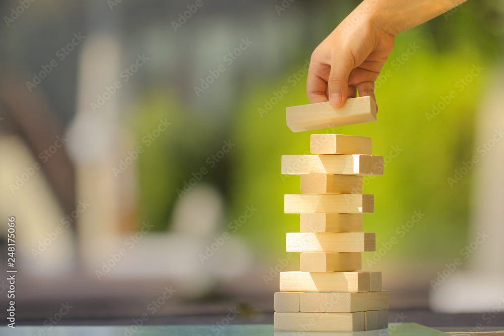Kid's hand holding wooden block to putting on wood stack tower game on greenery blurred background. Playing, learning, thinking, and planning concept Kid's hand holding wooden block to putting on wood stack tower game on greenery blurred background. Playing, learning, thinking, and planning concept