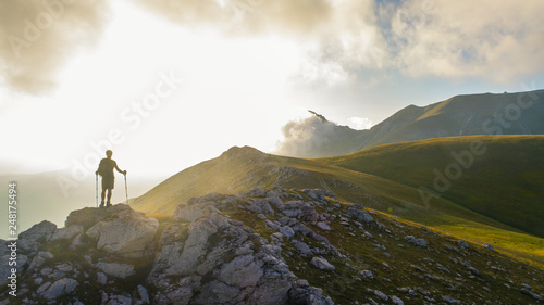 Young hiker man with backpack and walking poles, standing on peak of a mountain looking at sunset in cloudy sky. Green field and rocks. Abruzzo, Italy.