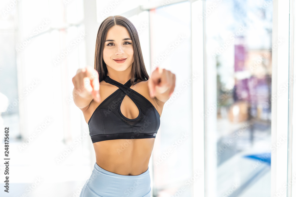 Photo of beautiful sports woman trainer standing and posing in gym while pointing and looking at camera.
