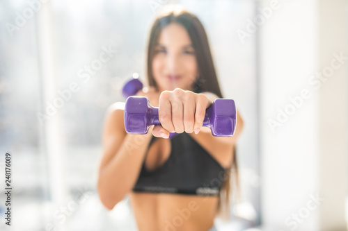 Young athletic woman pumping up muscles with dumbbells at fitness club in front of windows