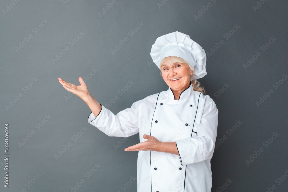 Senior woman chef studio standing isolated on gray pointing aside with hands on space looking camera joyful Senior woman chef studio standing isolated on gray pointing aside with hands on space looking camera joyful