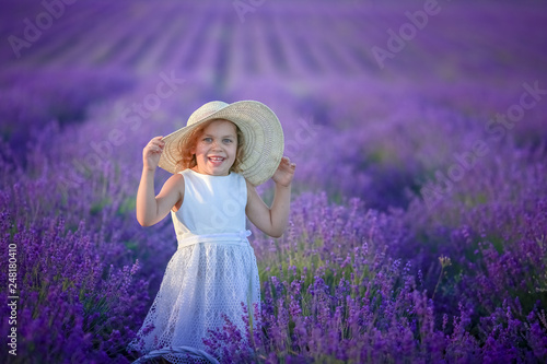 Cute young girl walking on a lavender field in white dress and hat on her head. Cute child face and nice hair with lavender bouquet 