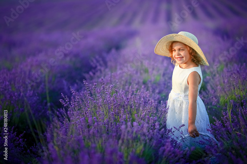 Cute curly young girl standing on a lavender field in white dress and hat with cute face and nice hair with lavender bouquet and smiling.