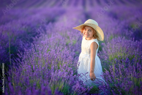 Cute young girl walking on a lavender field in white dress and hat on her head. Cute child face and nice hair with lavender bouquet 