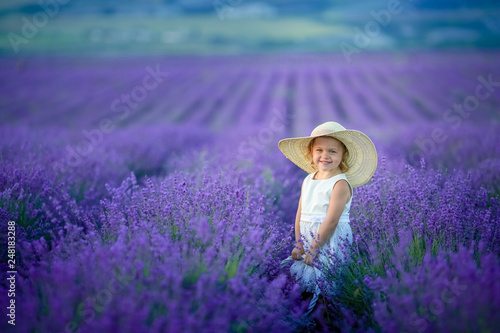 Cute curly young girl standing on a lavender field in white dress and hat with cute face and nice hair with lavender bouquet and smiling.