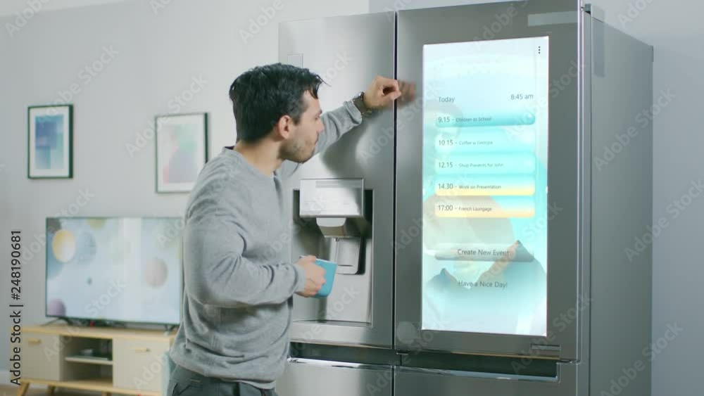 Handsome Young Man Walks Over to a Refrigerator While Drinking His Morning Coffee. He is Checking a To Do List on a Smart Fridge at Home. Kitchen is Bright and Cozy. Handsome Young Man Walks Over to a Refrigerator While Drinking His Morning Coffee. He is Checking a To Do List on a Smart Fridge at Home. Kitchen is Bright and Cozy.