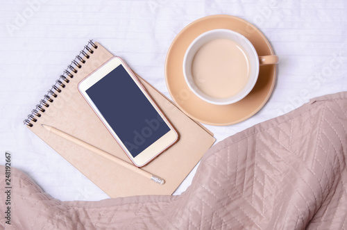 Cup of coffee with saucer notepad pencil smartphone on a white bed brown blanket home design, top view