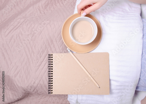 Woman in the white bed cup of coffee in hand notebook pencil brown blanket, top view