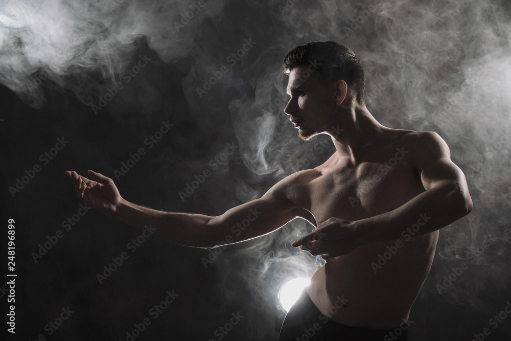 A young male ballet dancer with black leggings and a naked torso performs dance moves against a gray grunge background, with a light of lights and smoke.