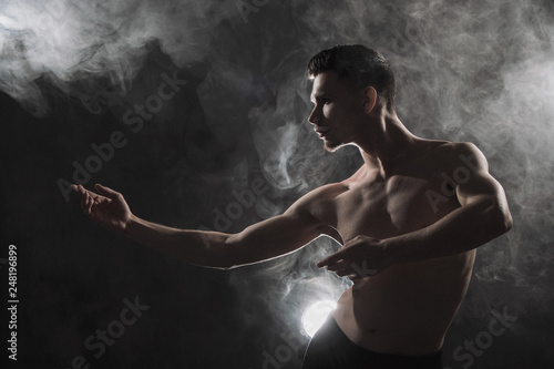 A young male ballet dancer with black leggings and a naked torso performs dance moves against a gray grunge background, with a light of lights and smoke. © spaskov