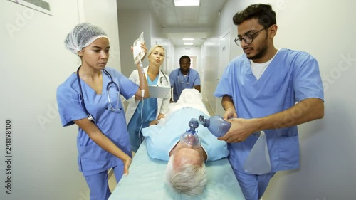 Dolly shot of blond female doctor giving instructions to group of ER nurses in scrubs administering oxygen and IV fluids to patient and rushing him to surgery