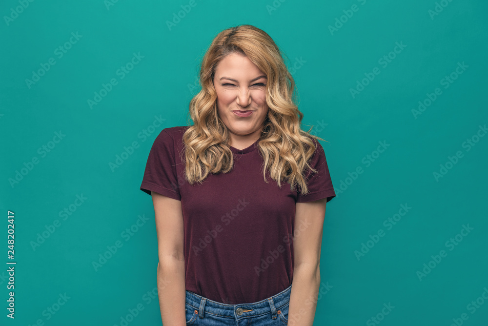 A young woman is squinting a sour grimace while standing against a blue background.