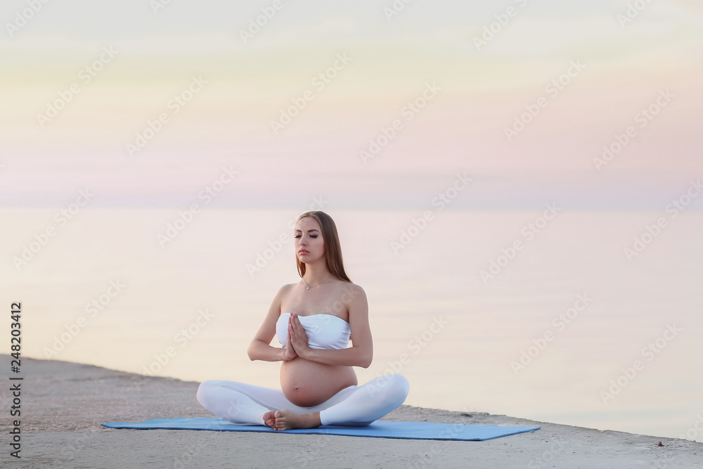 Young pregnant woman in white dress sitting on the beach near blue sea and breathing. Summer vacation during pregnancy, happy motherhood concept, close up on hands